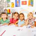 Czwórka dzieci w przedszkolu rysująca litery na kartkach papieru i patrząca w obiektyw aparatu.